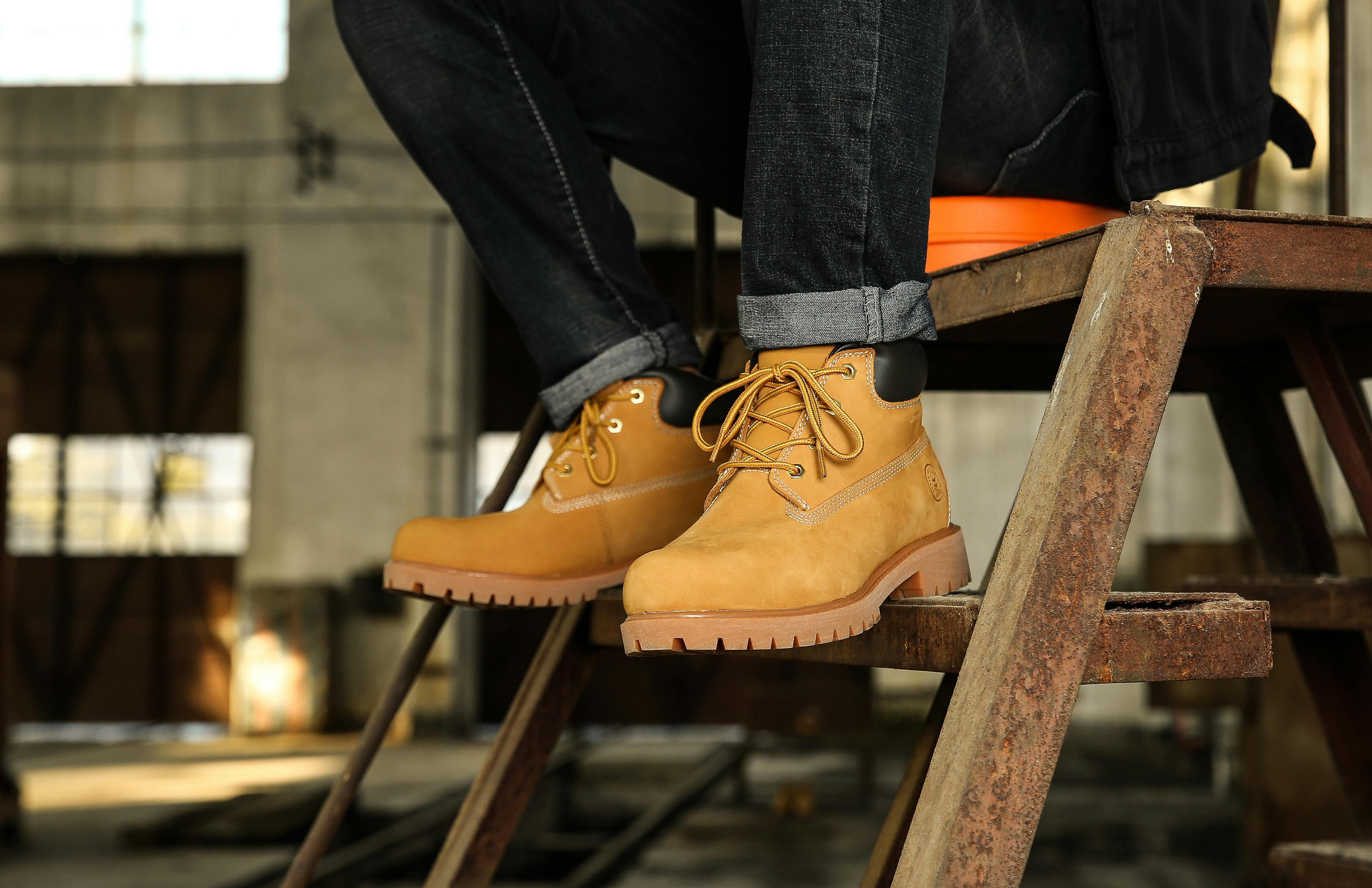 Close-up of a man's legs in jeans and brown hiking boots sitting on a metal ladder in an industrial